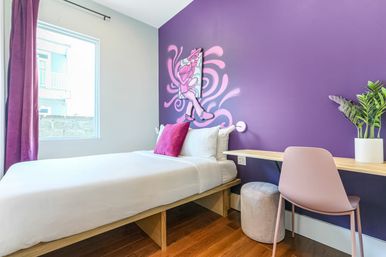 Bright modern bedroom with purple accent wall and pink mural, white bed with magenta pillow, light-wood desk, blush chair, potted plant, and sunlit window with purple curtains.