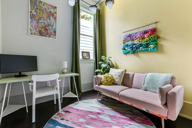 Vibrant, cozy apartment living room and home-office nook with a pink velvet sofa, patterned pillows and throw, mint-green desk with monitor and white chair, colorful braided wall hanging, potted plant and round multicolor rug.