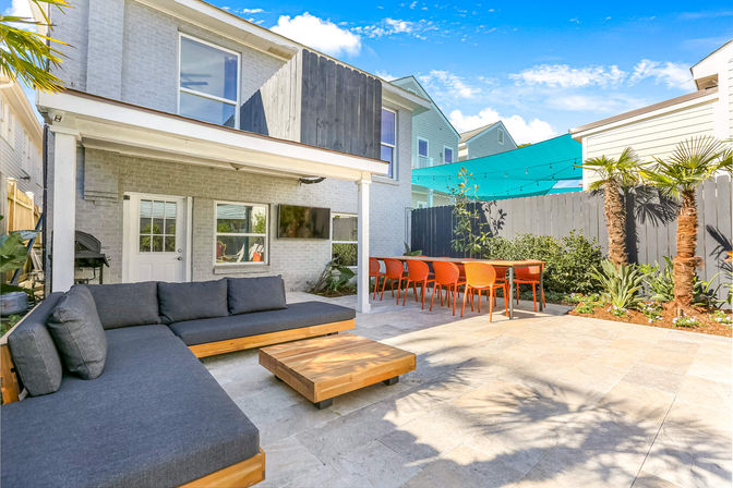 Sunny modern backyard patio with light-gray brick two-story house, covered porch, gray L-shaped outdoor sectional, low wooden coffee table, long dining table with bright orange chairs under a teal shade sail, potted palms and wall-mounted TV.