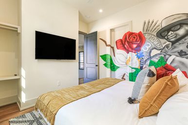 Bright modern bedroom with colorful floral mural featuring a stylized grayscale figure in sunglasses, wall-mounted flat-screen TV, cozy bed with mustard throw and decorative pillows, and hardwood floors.