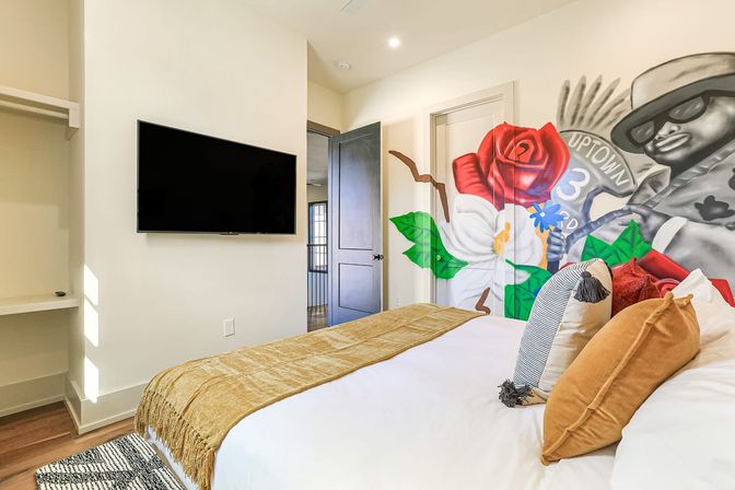 Bright modern bedroom with colorful floral mural featuring a stylized grayscale figure in sunglasses, wall-mounted flat-screen TV, cozy bed with mustard throw and decorative pillows, and hardwood floors.