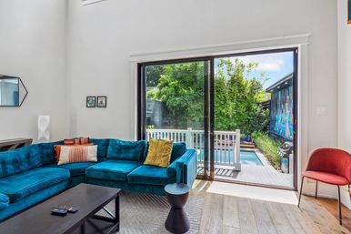 Bright modern living room with teal velvet sectional and wood floors, sliding glass doors opening to a fenced deck with a small plunge pool, bamboo greenery and a colorful mural outside.