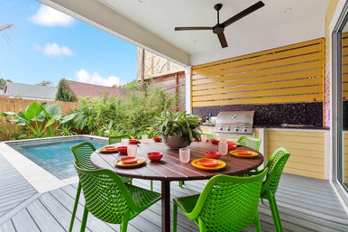 Covered backyard patio and outdoor dining area with a round wood table set with colorful plates and bowls, bright green chairs, potted centerpiece, ceiling fan, built-in stainless steel grill, and a swimming pool surrounded by lush tropical landscaping.