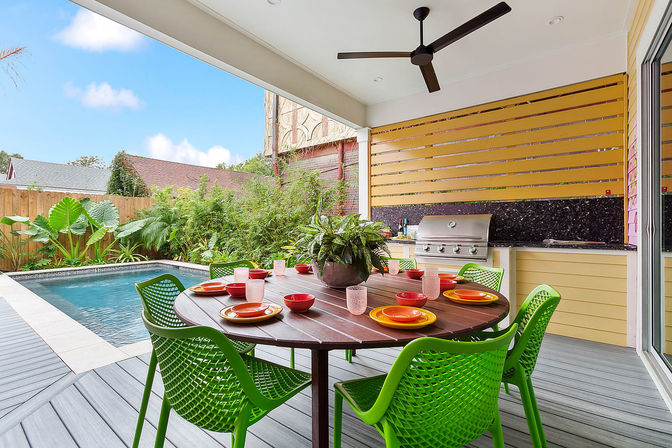 Covered backyard patio and outdoor dining area with a round wood table set with colorful plates and bowls, bright green chairs, potted centerpiece, ceiling fan, built-in stainless steel grill, and a swimming pool surrounded by lush tropical landscaping.