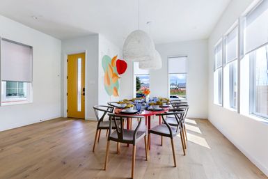 Sunlit modern dining room with a round red table set for eight, gray wood-frame chairs, woven white pendant lights, colorful wall art, a yellow front door, large windows and light oak floors.