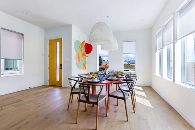 Sunlit modern dining room with a round red table set for eight, gray wood-frame chairs, woven white pendant lights, colorful wall art, a yellow front door, large windows and light oak floors.