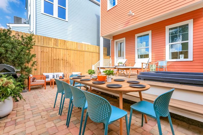 Bright coastal-style backyard patio with a wooden dining table set for eight, turquoise perforated chairs, brick paver floor, coral-colored house siding, wooden privacy fence and cozy lounge seating.
