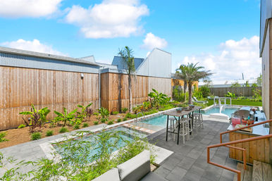 Sunny modern suburban backyard with narrow lap pool and spa, palm trees and tropical landscaping, bar-height table with stools, lounge seating and wooden privacy fence on a paved patio.