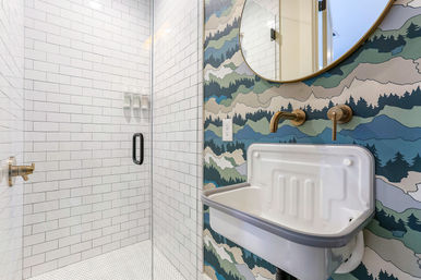 Modern bathroom with glass walk-in shower and white subway tile, brass wall-mounted faucet and round mirror over a white wall-mounted sink on mountain-pattern wallpaper