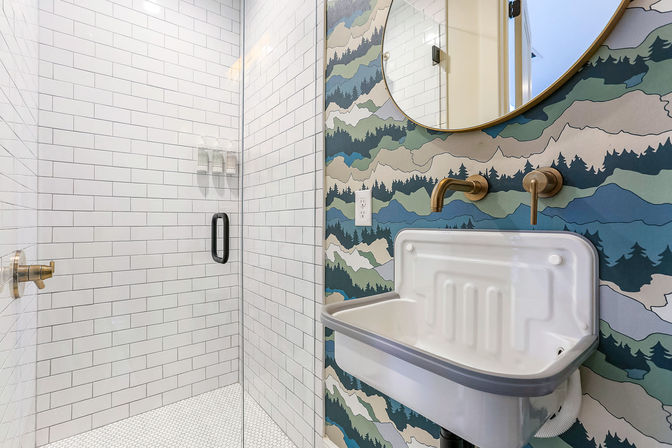 Modern bathroom with glass walk-in shower and white subway tile, brass wall-mounted faucet and round mirror over a white wall-mounted sink on mountain-pattern wallpaper