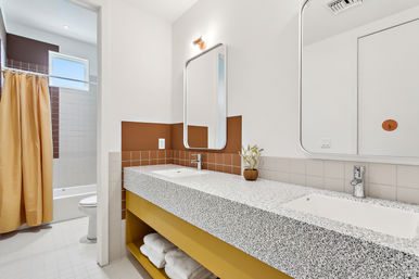 Bright modern bathroom with terrazzo double-sink vanity, mustard-yellow open shelf, brown tiled backsplash, rounded mirrors, and a white-tiled shower with beige curtain