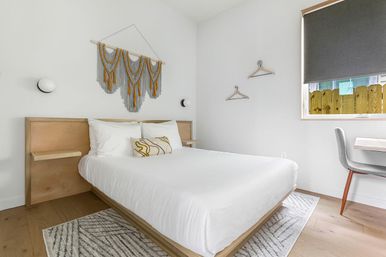 Bright modern minimalist bedroom with a white platform bed, natural wood headboard and floating shelves, gray-and-mustard macramé wall hanging, patterned rug, window with dark blind, and a small desk with a gray chair.