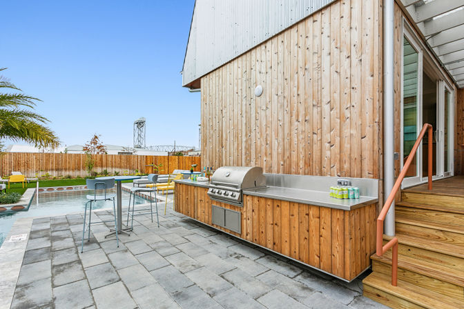 Sunny backyard patio with built-in stainless-steel grill set in a wooden outdoor kitchen, stone pavers, bar-height stools, poolside loungers and a rectangular swimming pool under a clear blue sky.