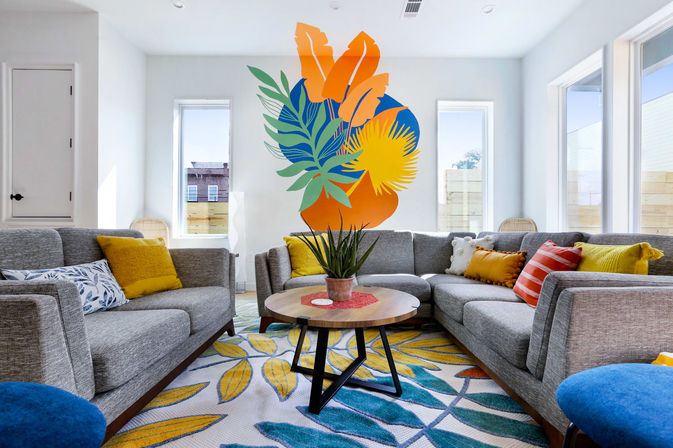 Sunlit modern living room with a gray sectional sofa, bold tropical wall mural, round wooden coffee table with potted plant, colorful leaf-pattern rug and yellow-orange throw pillows.