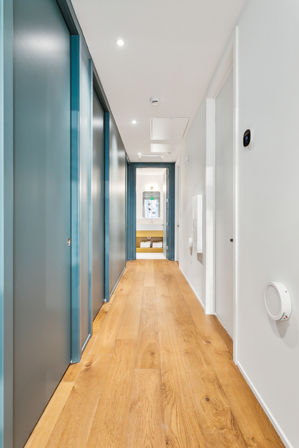 Sleek modern apartment hallway with light oak floors, teal sliding doors on the left, white walls, recessed ceiling lights, a round wall thermostat, and a bathroom with mirror and folded towels visible at the end.