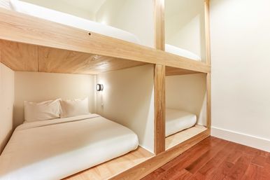 Cozy minimalist wooden bunk bed pods with crisp white linens, wall reading light, and warm hardwood floor in a capsule-style room.