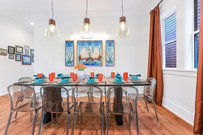 Bright modern dining room with three glass pendant lights, transparent acrylic chairs, hardwood floor, teal place settings and orange cups on a long table beneath coastal bird artwork and rust-orange curtains.
