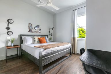 Cozy, light-filled modern bedroom with gray walls, dark wood bed and hardwood floors, white linens and orange accent pillow, decorative bird wall art, ceiling fan and a desk by a window with a leafy view.