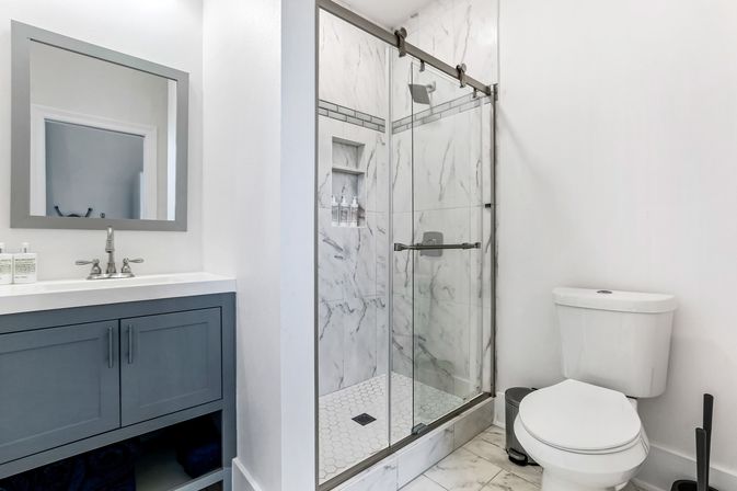 Bright modern bathroom with gray vanity, marble-tiled walk-in shower with sliding glass doors, hexagon tile floor, and white toilet