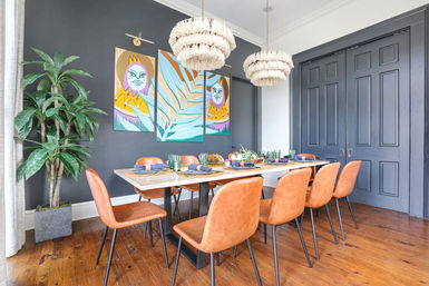 Modern dining room with eight orange leather chairs around a wooden table set for dinner, a black accent wall with a colorful botanical triptych, two tasseled pendant lights, large potted plant and dark paneled doors.