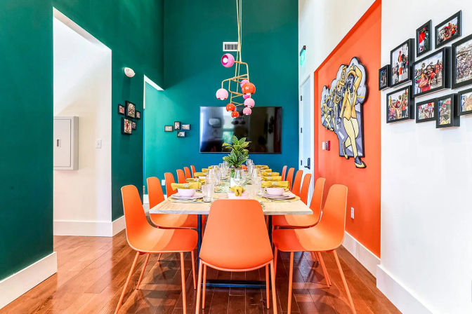 Vibrant contemporary dining room interior with a long set table, multiple orange chairs, teal and orange accent walls, colorful pendant globe lights, framed wall art and hardwood floors.