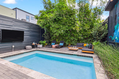 Modern urban backyard with a rectangular plunge pool, gray wood deck, wall-mounted TV, lush bamboo privacy hedge, lounge chairs and an inviting cushioned sectional with colorful pillows.
