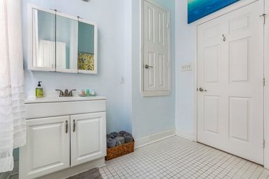 Sunlit pale-blue bathroom with white vanity and mirrored medicine cabinet, chrome faucet, woven basket of rolled gray towels, paneled white door and small square tiled floor