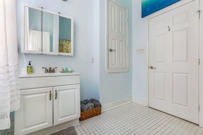 Sunlit pale-blue bathroom with white vanity and mirrored medicine cabinet, chrome faucet, woven basket of rolled gray towels, paneled white door and small square tiled floor