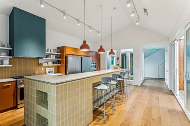 Bright modern open-plan kitchen with vaulted ceiling, long pale-tiled island and white countertop with four metal barstools, three terracotta pendant lights, stainless steel appliances, teal range hood, open shelving and warm wood floors