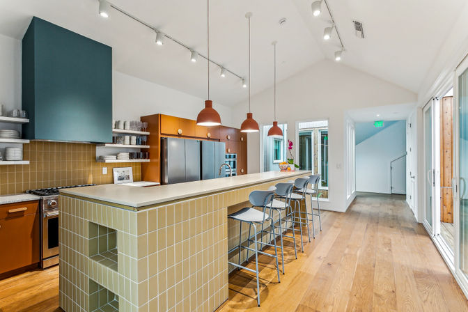Bright modern open-plan kitchen with vaulted ceiling, long pale-tiled island and white countertop with four metal barstools, three terracotta pendant lights, stainless steel appliances, teal range hood, open shelving and warm wood floors
