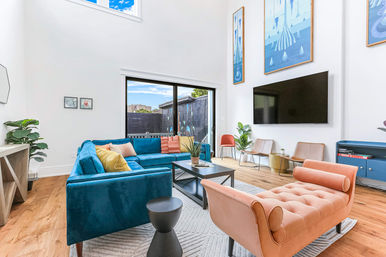 Sunlit double-height modern living room with a blue velvet sectional, peach tufted chaise, hardwood floors, large wall-mounted TV, sliding glass door to a patio, colorful wall art and potted plants — playful pops of color and contemporary style.