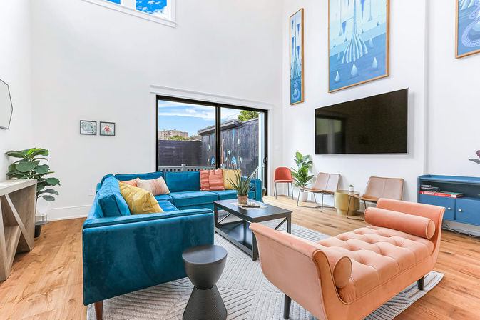 Sunlit double-height modern living room with a blue velvet sectional, peach tufted chaise, hardwood floors, large wall-mounted TV, sliding glass door to a patio, colorful wall art and potted plants — playful pops of color and contemporary style.