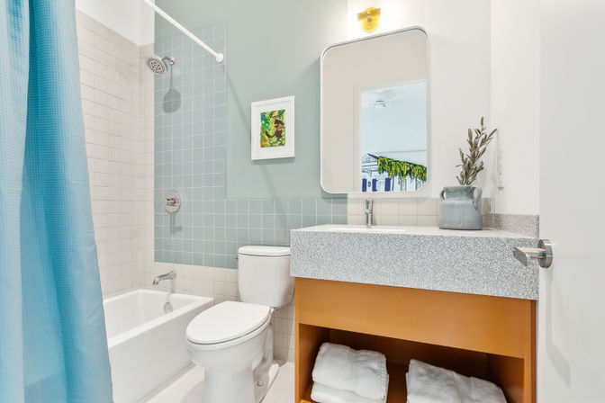 Bright modern bathroom with pastel blue square tiles, bathtub-shower with blue curtain, white toilet, speckled vanity countertop, mirror with sconce, open shelf holding folded towels and a ceramic vase.