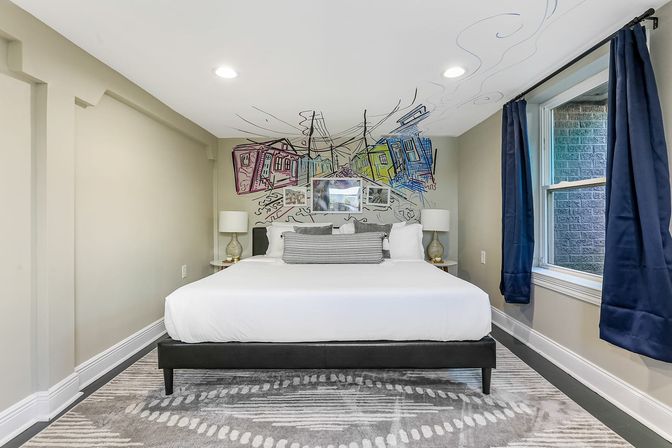 Modern bedroom with low ceiling, king bed in white linens, colorful urban sketch mural above the headboard, matching bedside lamps, navy curtains and a patterned gray area rug.