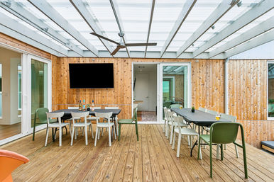 Covered modern backyard patio with wooden deck and cedar-clad walls, transparent pergola roof, two long dining tables with mixed white and green chairs, ceiling fan, wall-mounted TV and sliding glass doors.