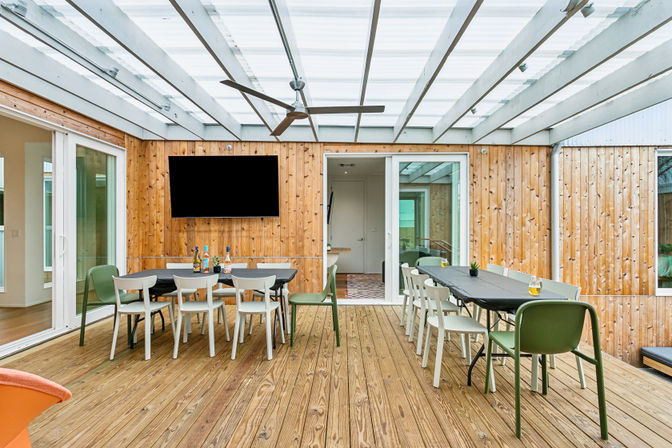 Covered modern backyard patio with wooden deck and cedar-clad walls, transparent pergola roof, two long dining tables with mixed white and green chairs, ceiling fan, wall-mounted TV and sliding glass doors.