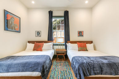 Cheerful modern guest room with two matching twin beds, wooden headboards, white linens, navy throws, orange accent pillows, mid-century nightstand and lamp under a curtained window, colorful striped rug and framed artwork.