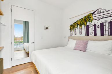 Bright modern minimalist bedroom with white linens and lavender accent pillow, hardwood floors, wall mural of a house topped with hanging yellow-green foliage, and a full-length mirror on the door reflecting a sunlit balcony and outdoor view.