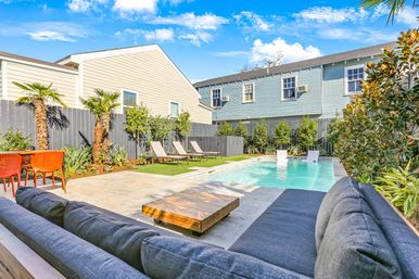 Sunny suburban backyard pool and patio with modern gray outdoor sofa and low wooden coffee table in foreground, rectangular swimming pool with in-pool loungers, three sunbeds on turf, orange dining chairs, palm trees and two-story homes behind a gray fence.