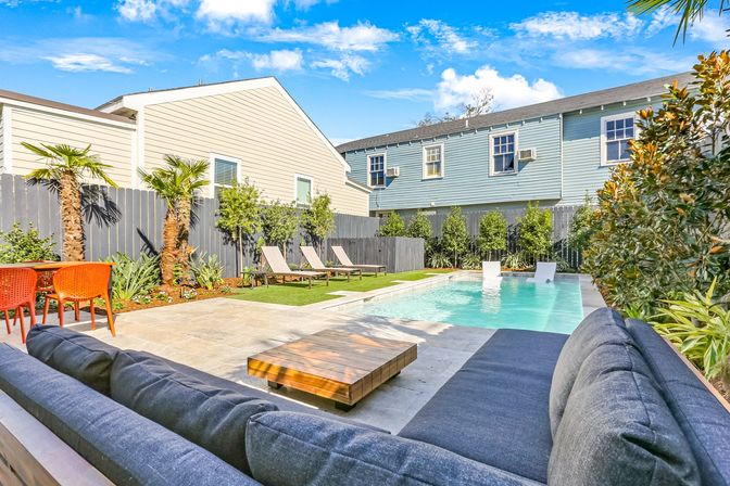 Sunny suburban backyard pool and patio with modern gray outdoor sofa and low wooden coffee table in foreground, rectangular swimming pool with in-pool loungers, three sunbeds on turf, orange dining chairs, palm trees and two-story homes behind a gray fence.