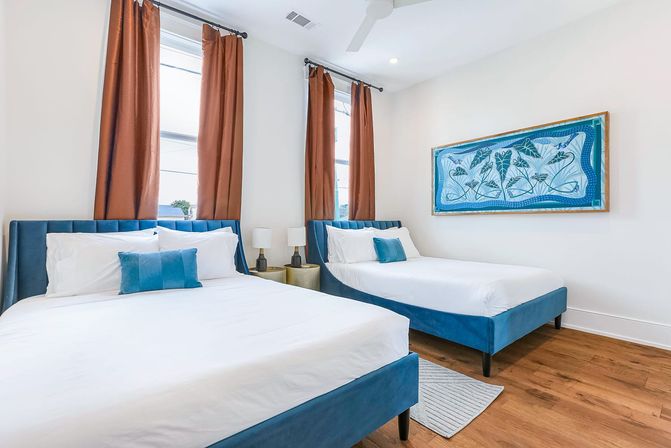 Bright modern guest bedroom with two blue-upholstered beds, crisp white linens and teal throw pillows, hardwood floors, rust-colored curtains, and framed blue botanical artwork.