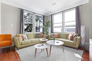 Sunlit modern living room with olive-green mid-century sofas in an L-shape, round white nesting coffee tables, patterned rug, large windows with gray curtains, orange accent chair, tall potted plant and hardwood floors.