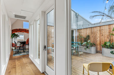Bright modern apartment hallway with hardwood floors and glass door opening to a sunny wooden patio deck featuring corrugated planter boxes, string lights, turquoise bistro set and a yellow chair; indoor dining nook with patterned accent wall visible beyond.