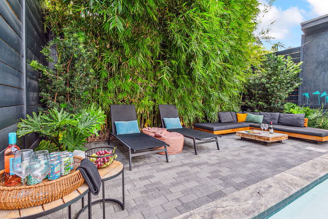 Inviting modern poolside patio with two black lounge chairs and blue pillows, L-shaped gray outdoor sofa with colorful throw pillows, low wooden coffee table with wine and glasses, wicker tray of drinks and fruit, and tall bamboo privacy screen.