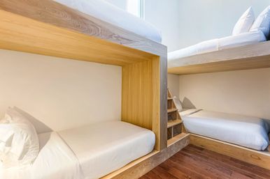 Bright modern bedroom featuring minimalist wooden built-in bunk beds with white linens, a small stepped ladder and warm hardwood floors.