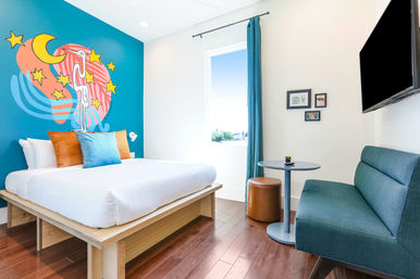 Bright modern hotel room with king bed, teal accent wall featuring a whimsical moon-and-stars mural, teal sofa, round table, wall-mounted TV and hardwood floor