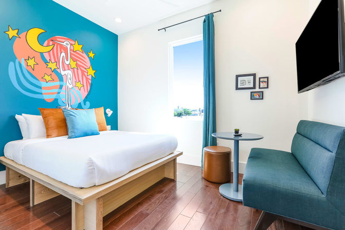 Bright modern hotel room with king bed, teal accent wall featuring a whimsical moon-and-stars mural, teal sofa, round table, wall-mounted TV and hardwood floor