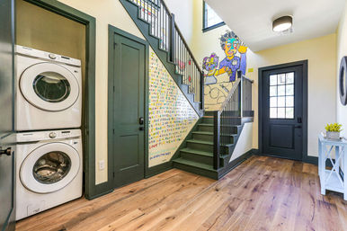 Bright modern entryway with stacked washer and dryer in closet, dark green stairs and trim, colorful mural under the staircase, wide hardwood floors and a black paneled door.