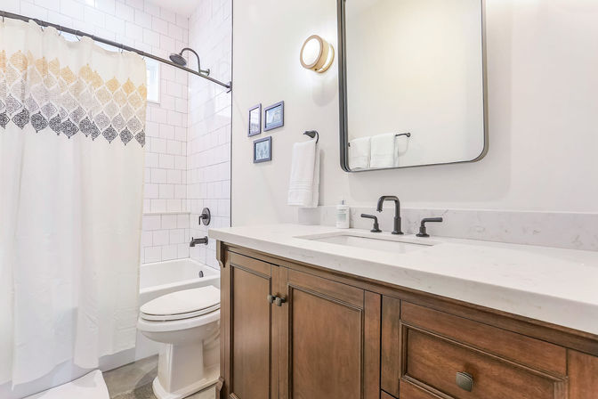 Bright modern bathroom with white subway-tiled tub-shower and patterned curtain, wood vanity with marble countertop, black matte faucet and hardware, rectangular mirror and round wall sconce