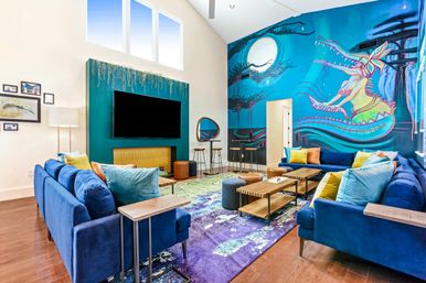 Bright modern living room with high ceilings, blue velvet sofas and colorful throw pillows, large flat-screen TV above a teal fireplace, wooden coffee tables on a purple patterned rug, and a vibrant blue wall mural featuring a moonlit scene and playful dragon-like creature.